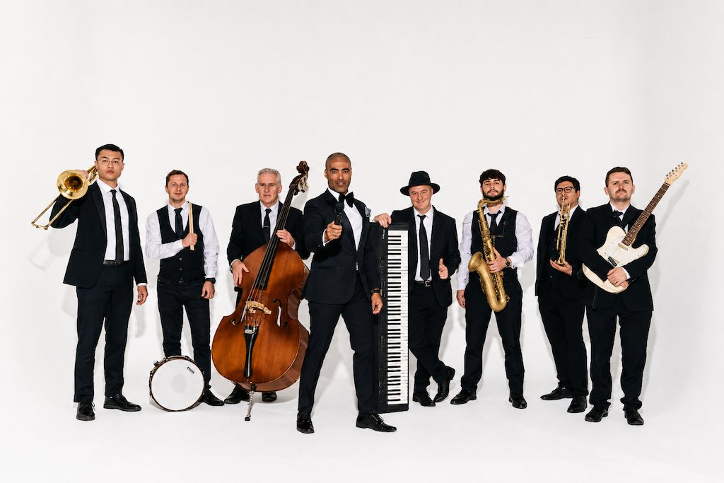 The eight members of the band in black suits with their instruments: trombone, drums, double bass, keyboard, saxophone, trumpet and guitar, with Luke in the centre holding a microphone