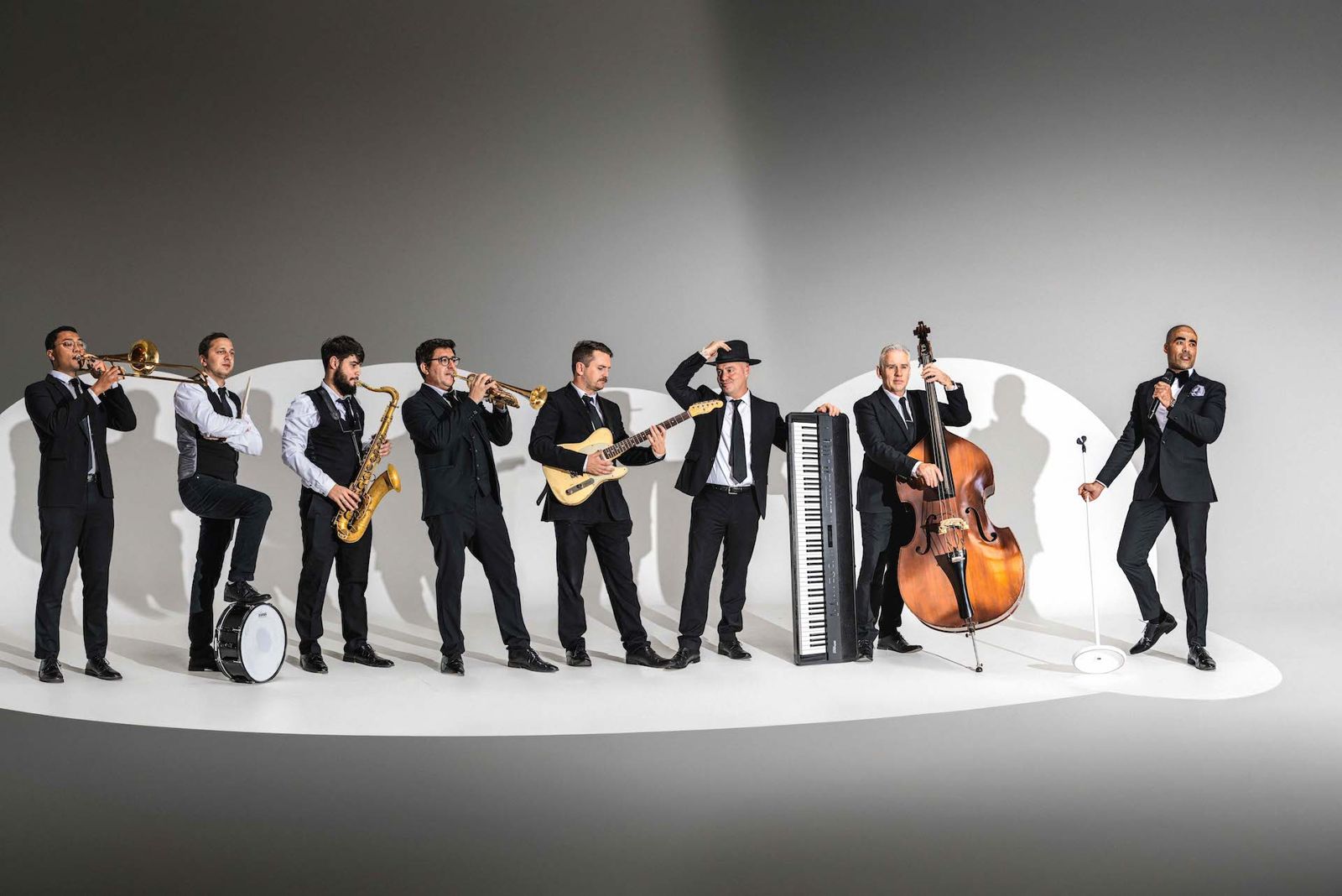 The band lined up in a spotlight with trombone, snare drum, saxophone, trumpet, guitar, keyboard and double bass, Luke singing at the end of the line