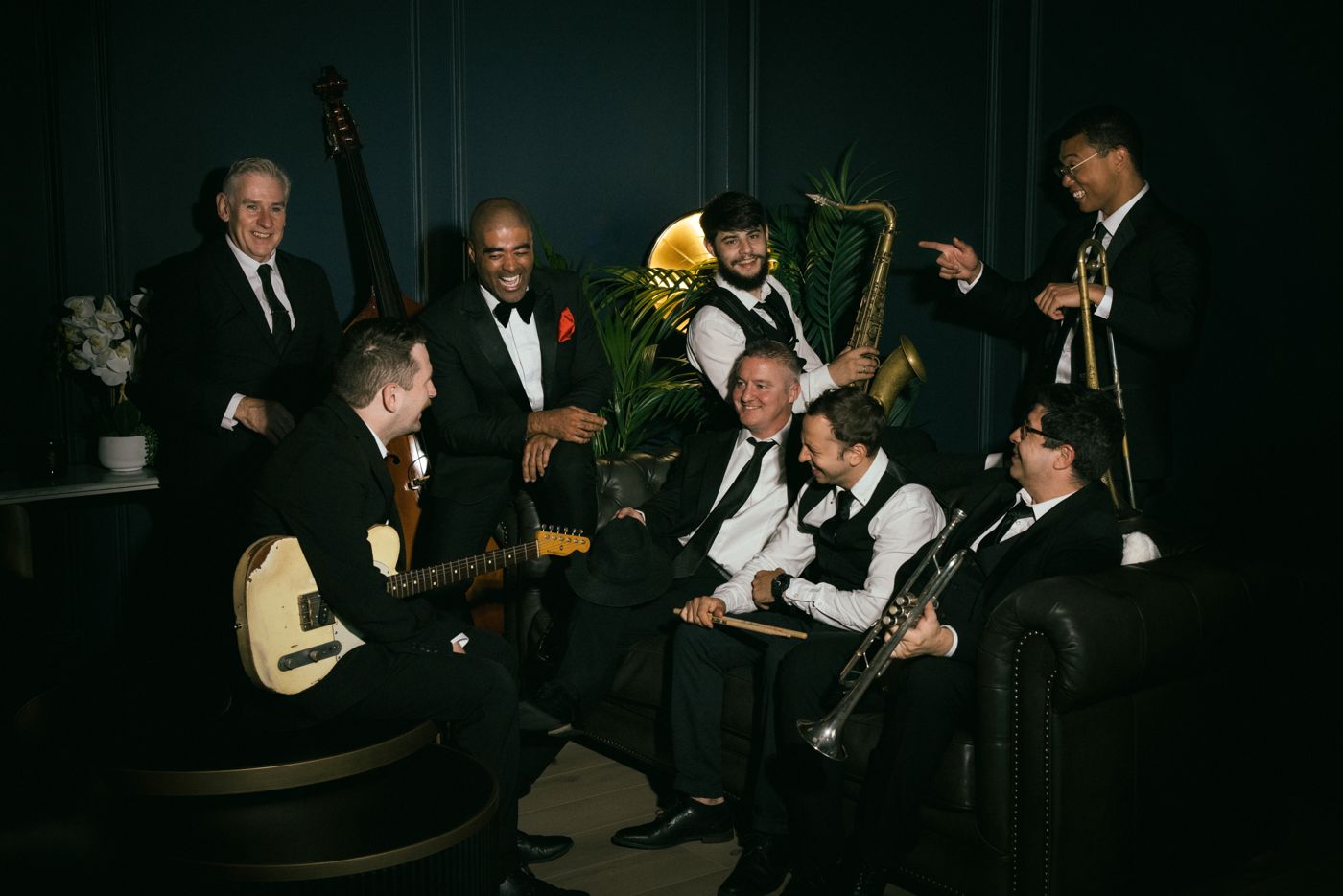 The band relaxing and laughing together on a leather sofa in a dark panelled lounge, holding their instruments