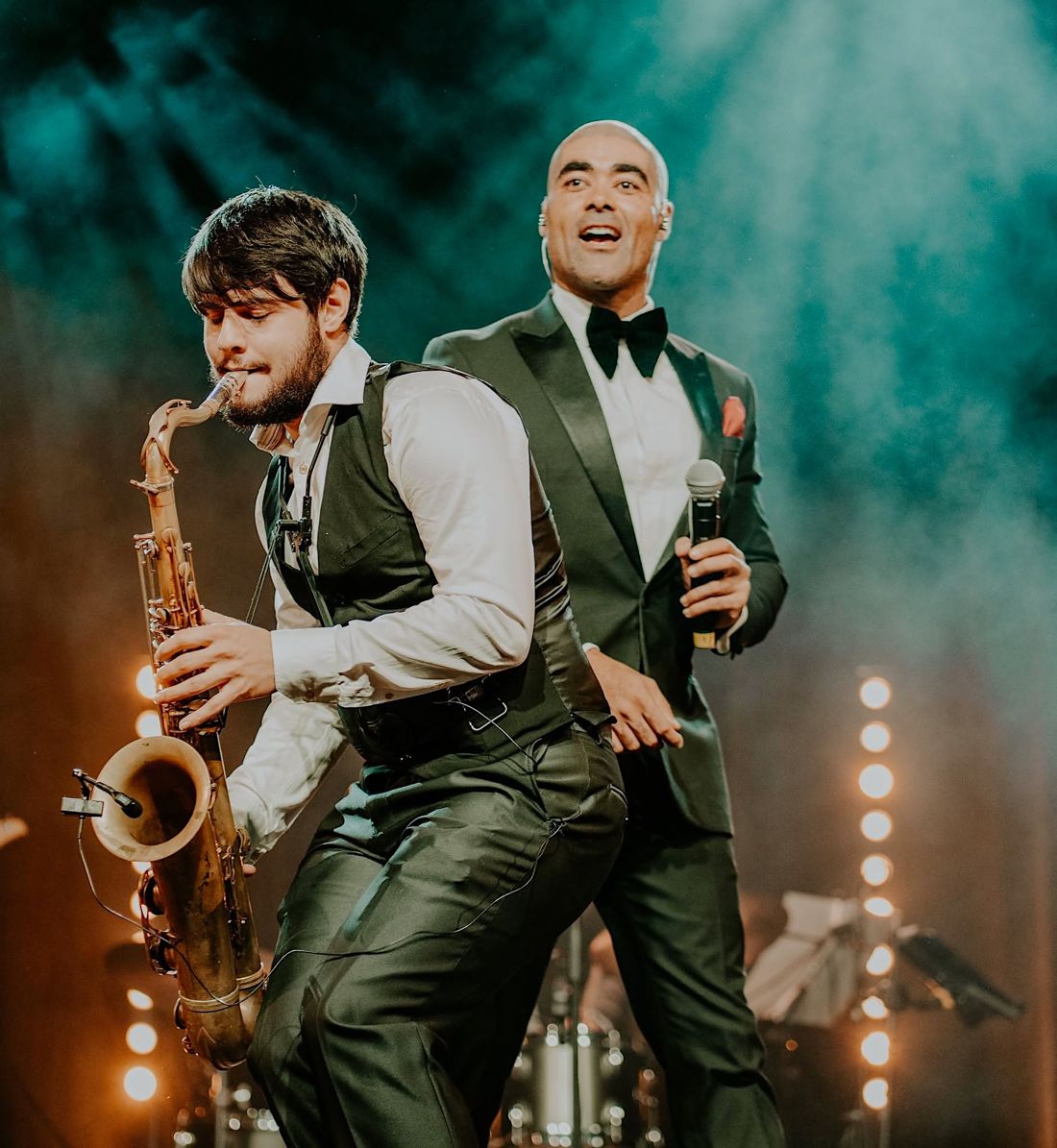 Luke Thomas singing on stage in a tuxedo behind the saxophonist mid-solo, under teal smoke and stage lights